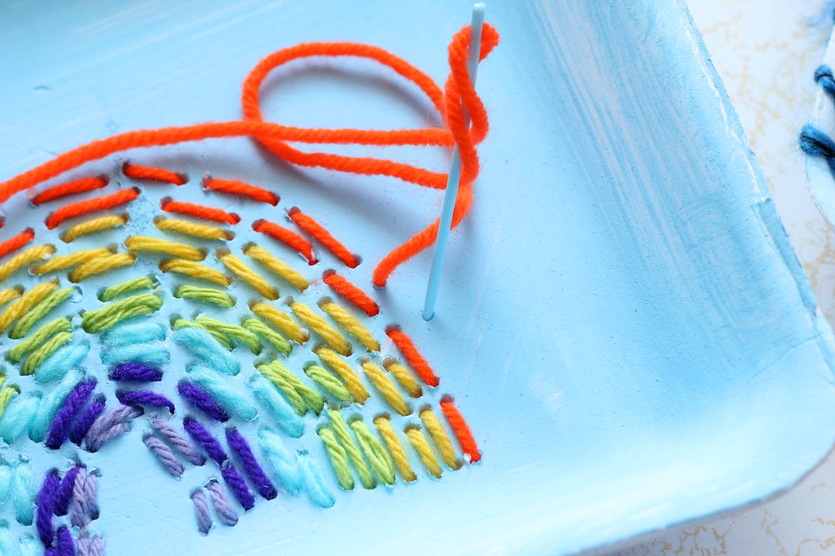 close up plastic sewing needle sewing orange band of rainbow on styrofoam tray